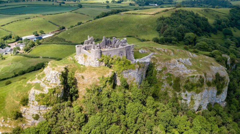 An aerial view of the historic stone ruins of Carreg Cennen Castle perched dramatically atop a steep, rugged limestone crag. The fortress overlooks a sprawling, lush green landscape of rolling hills, trees, and patchwork farm fields in Carmarthenshire under a bright sky.