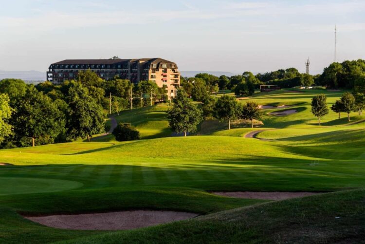A scenic view of the Celtic Manor Resort. In the foreground, a pristine, rolling green golf course features sand bunkers and scattered trees illuminated by warm sunlight, casting long shadows. In the background, the large, multi-story resort hotel building stands against a clear blue sky.