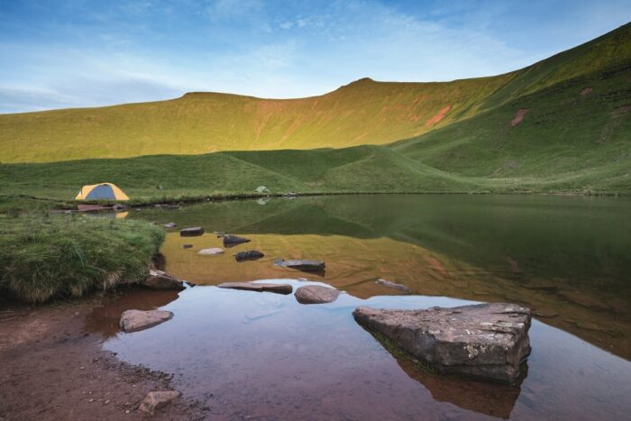 A yellow and grey tent is set up on grassy ground by a calm lake, with large rocks in the water and green hills with Pen y Fan in the Brecon Beacons, brightly lit by the setting sun. The sky is mostly clear with a few light clouds.