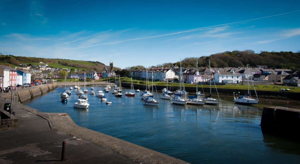 A view of the inner harbour at Aberaeron in Mid Wales, with several small sailboats and motorboats moored in calm water, surrounded by rows of brightly painted Georgian houses under a clear blue sky.