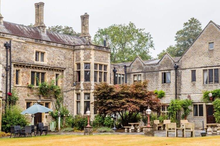 A large stone house with tall windows and ivy-covered walls, surrounded by trees. The courtyard features garden furniture, lamp posts, and a green lawn, creating a peaceful, historic setting.