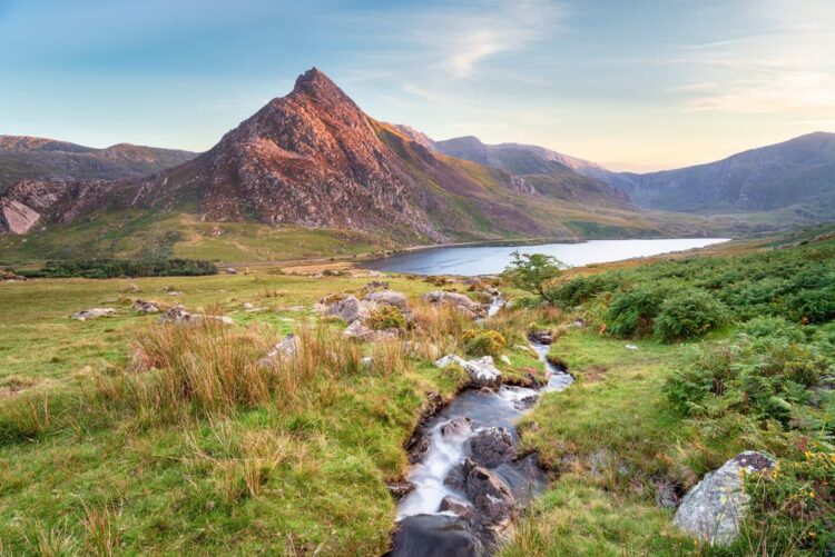 Mount Tryfan in the distance featuring a stream flowing through grassy fields with rocks, leading to a lake surrounded by mountains under a clear sky.