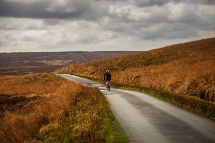 A lone cyclist rides on a narrow, winding road through hilly moorland with brown grass under a cloudy sky—a peaceful, remote scene capturing the essence of cycling in Wales.
