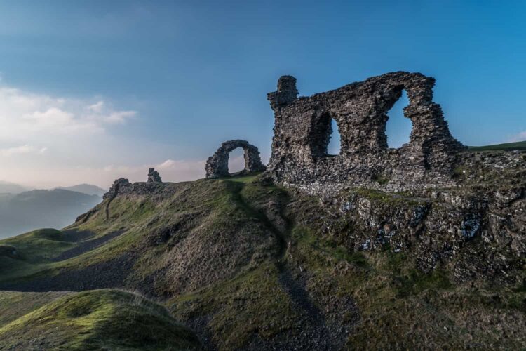 The ancient, jagged stone ruins of Castell Dinas perched high on a rugged, grassy hilltop in North West Wales. The crumbling walls feature large, arched openings and stand dramatically against a hazy, partly cloudy sky, with rolling green hills fading into the distant background.