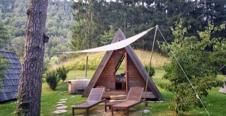 A small A-frame wooden cabin with an open front sits on grassy ground, shaded by a white tarpaulin—perfect for glamping in North Wales. Two wooden lounge chairs invite relaxation among the trees, with a forested hillside as the backdrop.