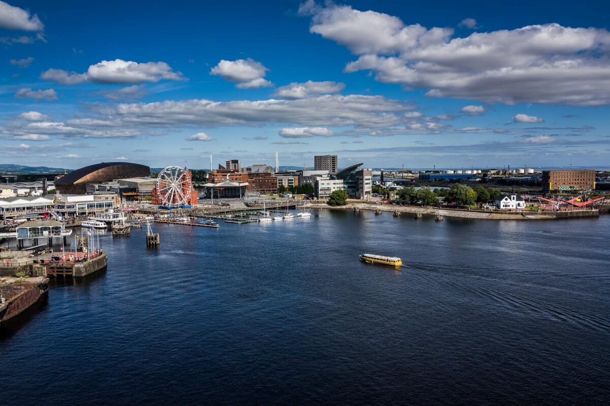 A bright, wide-angle view across the waters of Cardiff Bay on a sunny day. The waterfront skyline features the copper dome of the Wales Millennium Centre, a large Ferris wheel, the white Norwegian Church, and a yellow boat sailing across the bay, showcasing popular things to do in Cardiff.