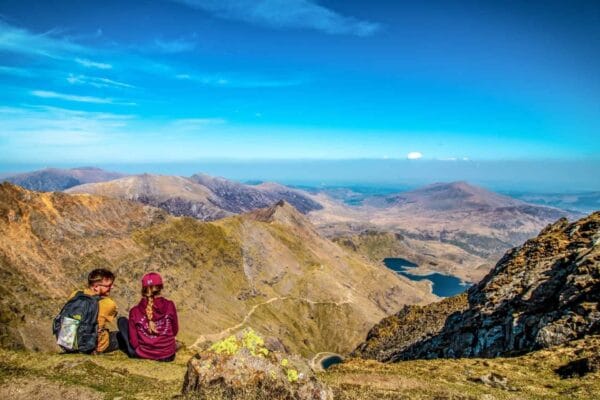 A man and woman sitting on the ground looking out to Eryri (Snowdonia) National Park with the hills and mountains clearly seen on a bright summer day