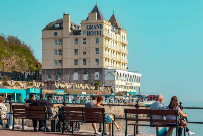 People sit on benches along a seaside promenade with the Grand Hotel—one of the best hotels in Wales—in the background under a clear blue sky. Some small shops and cafés are visible near the water.