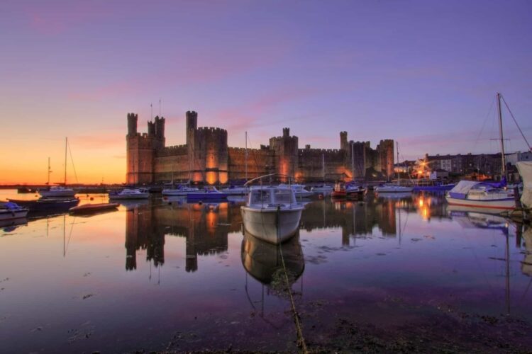 Caernarfon Castle’s historic stone towers are illuminated at sunset, reflected in calm harbour waters. Several boats are moored in the foreground, set against a vibrant purple and orange sky.