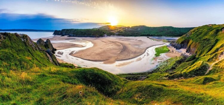 A panoramic view of Three Cliffs Bay -sandy beach with a winding river, surrounded by green cliffs and grassy hills, under a clear sky with the sun setting—perfect inspiration for Wales holiday ideas.