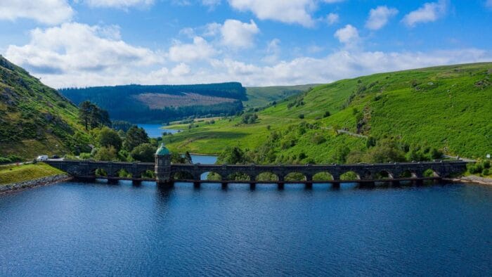 A stone dam with arches stretches across a deep blue Elan Valley reservoir, surrounded by green rolling hills and trees under a bright, partly cloudy sky. A small stone tower with a green dome sits on the dam.