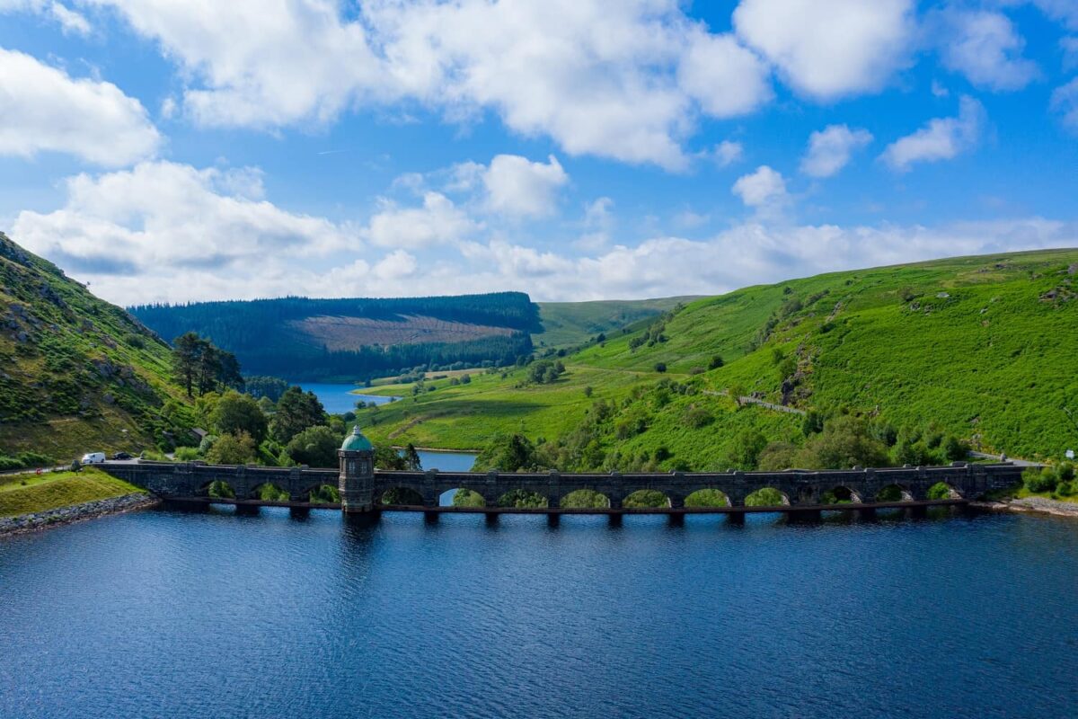 A wide view of the Craig Goch dam in the Elan Valley, featuring a long arched stone wall and a copper-domed tower spanning a deep blue reservoir, surrounded by bright green hills under a cloudy summer sky.