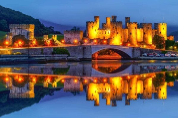 Conwy Castle lit up in yellow light at dusk with the bridge and river showing in the foreground