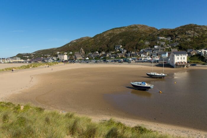 Two boats rest on the sandy shore at low tide, with grassy dunes in the foreground and a small coastal town nestled at the base of green hills—an idyllic scene typical of the best beaches in Wales under a clear blue sky.