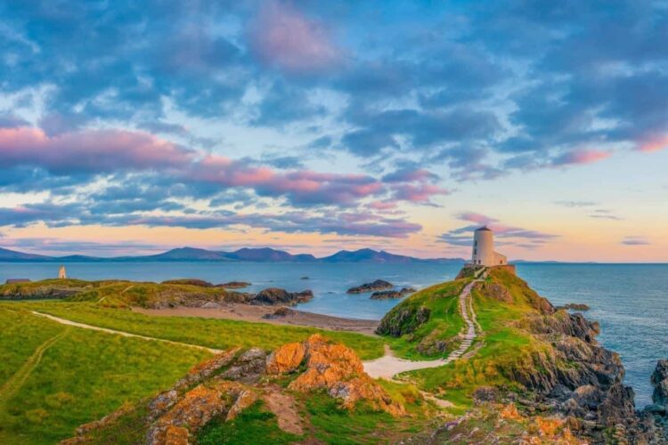 A coastal scene at sunset shows a winding path along the Wales Way leading to a white lighthouse on a rocky hill, surrounded by green grass and overlooking the sea, with colourful clouds in the sky.