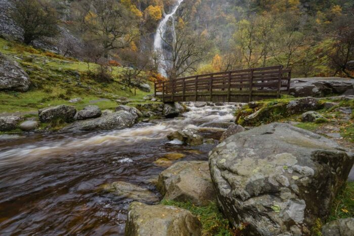 A wooden footbridge crosses a rocky stream surrounded by green grass and mossy boulders, with Aber Falls cascading down a forested hillside—a perfect scene on an Aber Falls day walk in North Wales.
