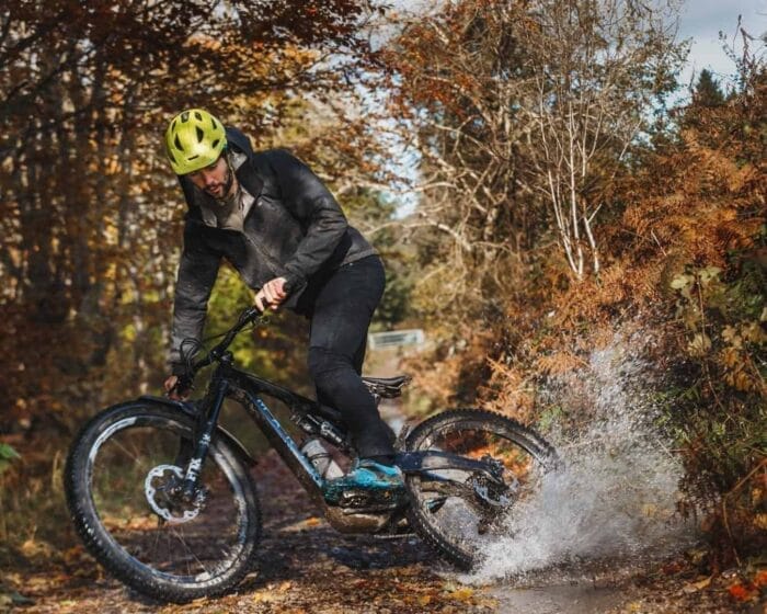 A person wearing a yellow helmet and dark clothing rides a mountain bike on a forest trail, splashing through a puddle amid vibrant autumn foliage