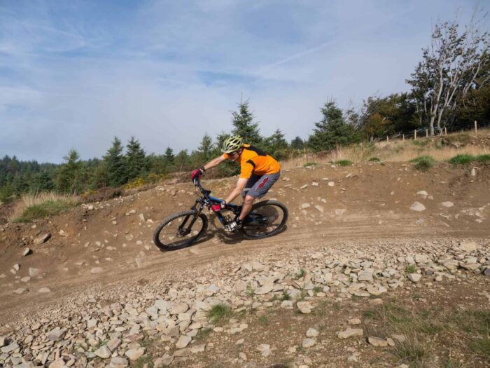 A person wearing an orange shirt and helmet rides a mountain bike on a rocky, dirt trail surrounded by grass and trees under a partly cloudy sky, capturing the spirit of Adventure in Wales.