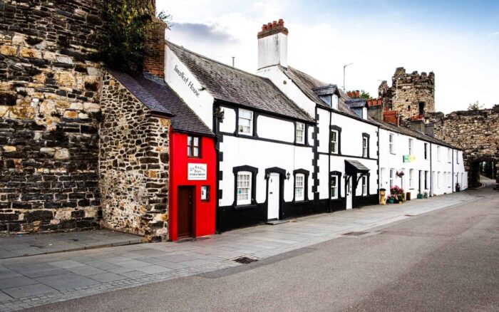 A row of quaint, historic cottages with white walls and black trim lines a quiet street; the world’s smallest house, painted bright red, stands out beside a stone castle wall under a partly cloudy sky.