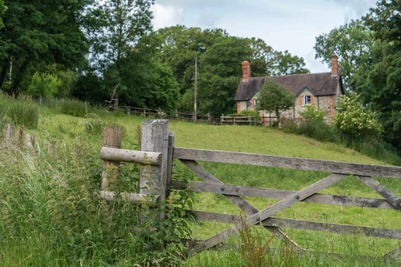 A wooden gate stands open in front of a grassy field, with a rustic brick house and tall trees in the background—perfect for those seeking holiday cottages in Pembrokeshire on a partly cloudy day.