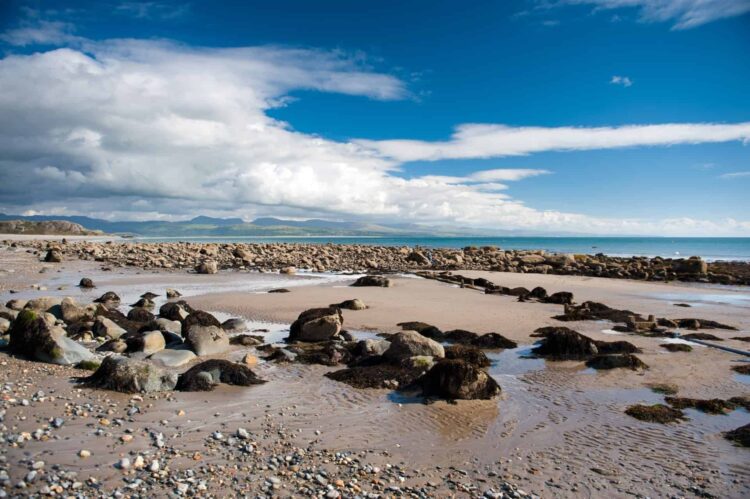 Criccieth Beach near Porthmadog at low tide with wet sand, scattered stones, and pools of water under a partly cloudy blue sky. Mountains and sea are visible in the distance, making it one of the best beaches in Wales to explore at sunrise or sunset.