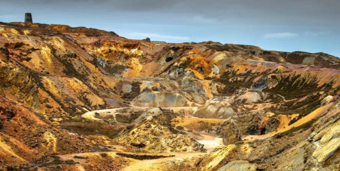 A rugged, colourful landscape featuring rocky hills in yellow, orange, and brown. Sparse vegetation dots the terrain, with a small old stone structure on a distant hill—one of the unique Isle of Anglesey tourist attractions under a cloudy sky.