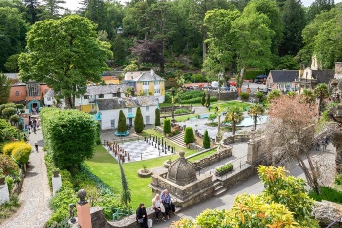A vibrant village, Portmeirion, with colourful buildings, manicured gardens, fountains, and a large chessboard surrounded by lush green trees, with people strolling along pathways