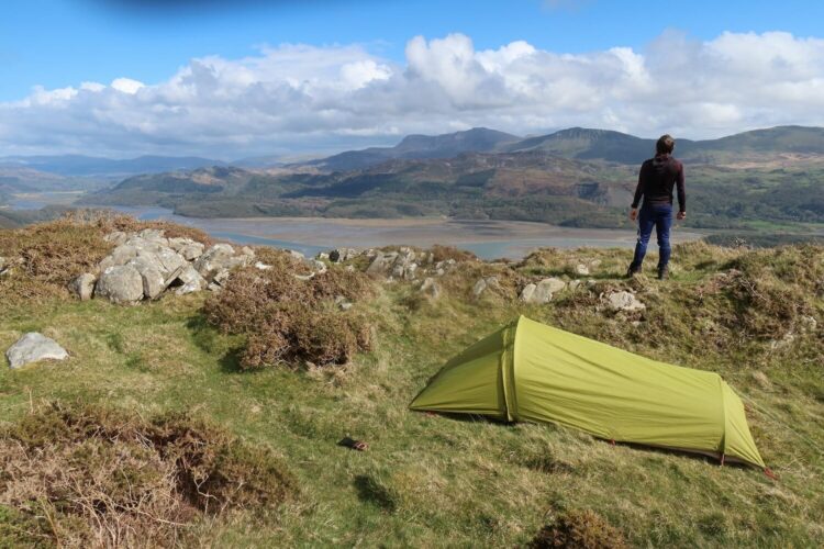 A person stands on a grassy hill overlooking a scenic valley with mountains, a river, and clouds. In the foreground, a small green tent is pitched near rocks and bushes, capturing the essence of wild camping in Wales.