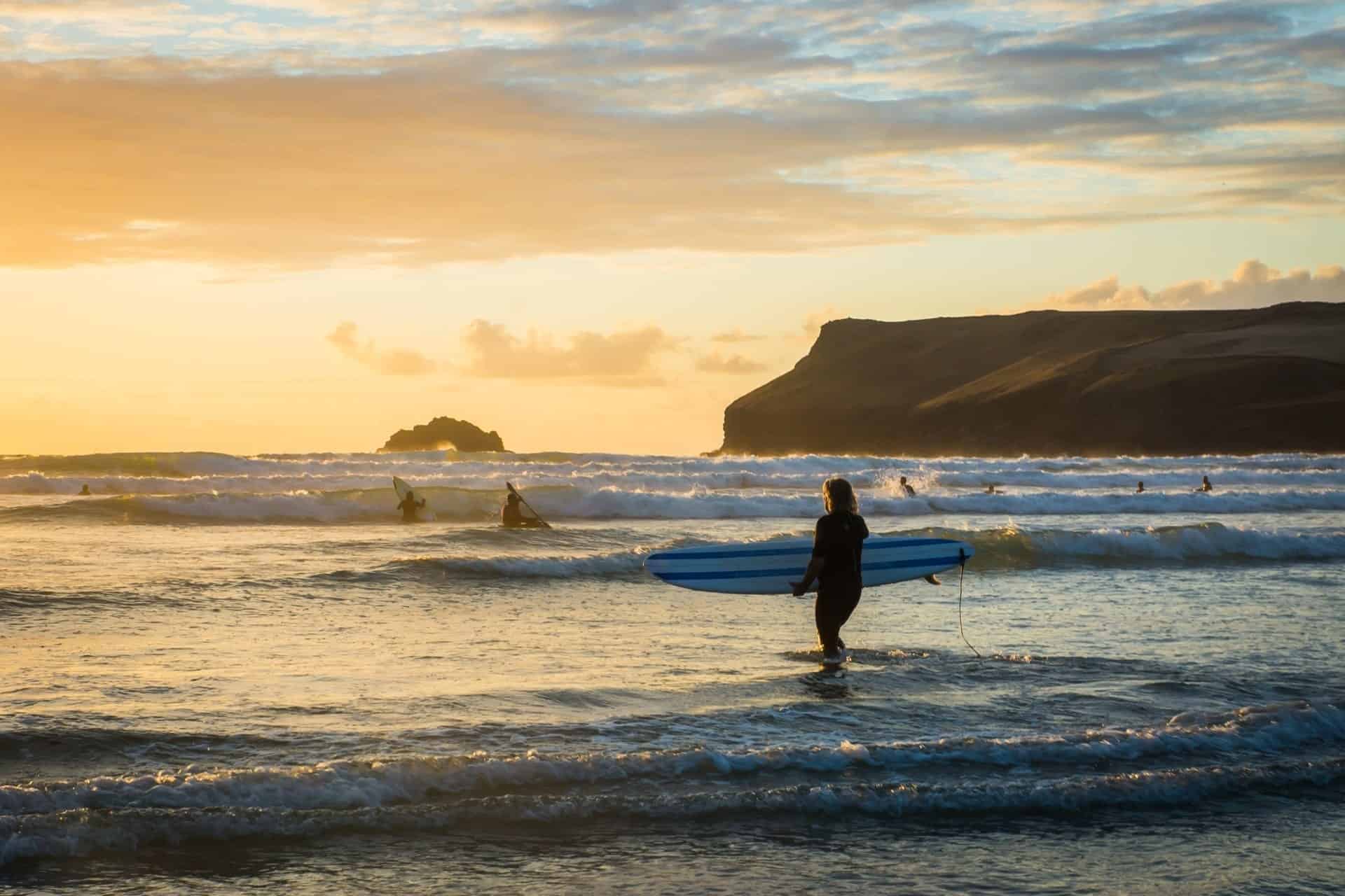 A person carrying a surfboard walks into the sea at sunset, with waves, surfers, and a rocky coastline in the background—capturing the beauty often seen at surfing beaches in Wales under a partly cloudy sky.