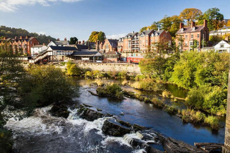 A picturesque view of the town of Llangollen in North East Wales. In the foreground, a river flows rapidly over a rocky bed, creating small white-water cascades. Along the riverbank, a historic railway station featuring a white footbridge and a red train carriage is nestled in front of charming multi-story brick buildings and lush, tree-covered hills beneath a bright blue sky.