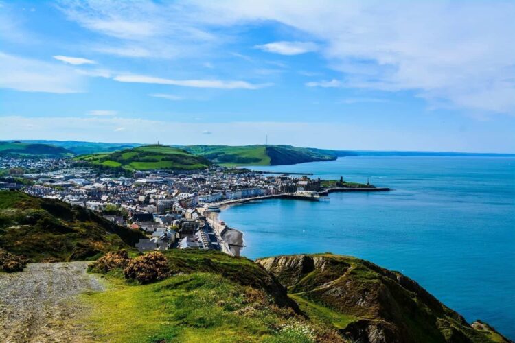 A sweeping, high-angle view overlooking a picturesque coastal town nestled along the bright blue waters of Cardigan Bay. In the foreground, a grassy green clifftop slopes down toward the curving shoreline, promenade, and dense buildings of the town. Rolling green hills rise in the background beneath a wide, sunny blue sky.
