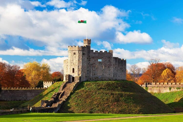 A stone castle keep stands atop a grassy hill, with stone steps leading up. The Welsh flag flies from the tower of Cardiff Castle. Trees with autumn leaves and a partly cloudy sky create a picturesque backdrop.