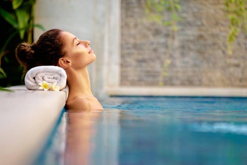 A woman relaxes in a pool at a spa hotel in Wales, leaning against the edge with her eyes closed. Her head rests on a rolled towel with a frangipani flower, surrounded by lush green plants and a stone wall in the background.