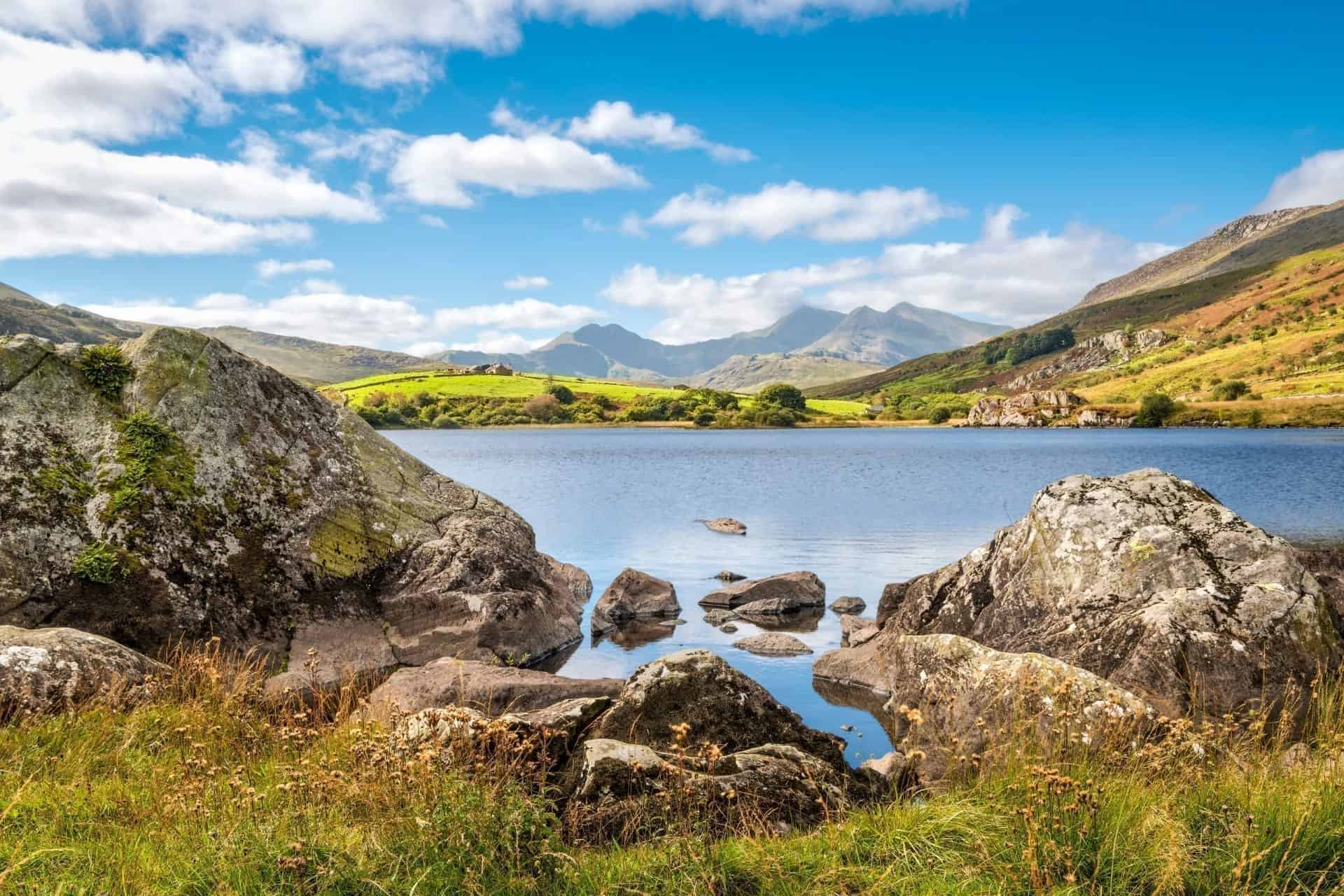 A picturesque landscape featuring large rocks on the grassy shore of a calm blue lake, looking towards the majestic peak of Mount Snowdon in the distance under a bright blue sky. Exploring these scenic surrounding trails offers an unforgettable experience for horse riding in Snowdonia.