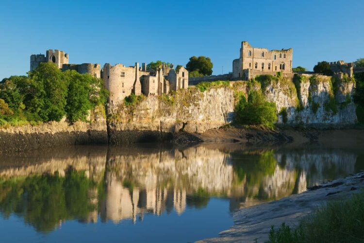 The historic stone ruins of Chepstow Castle perched atop a high, rocky limestone cliff overlooking a calm river. The water below creates a perfect mirror reflection of the majestic castle walls, sheer cliffs, and lush green trees under a bright, clear blue sky.