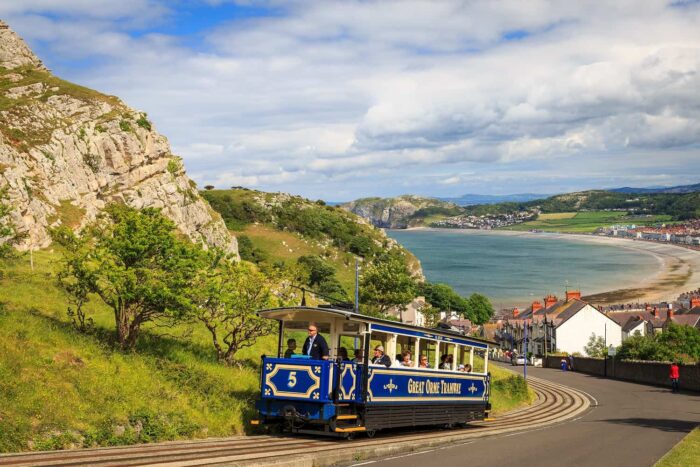 A blue and white vintage tram climbs a hillside track in Llandudno, overlooking the bay, sandy beach, and coastal town, with green hills and dramatic cliffs under a partly cloudy sky.