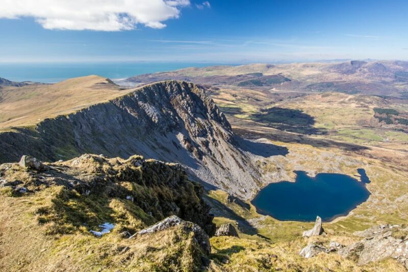 A view from Cadair Idris of a dramatic ridge overlooking a heart-shaped blue lake and vast green valleys under a bright, partly cloudy sky with distant rolling hills in the background.