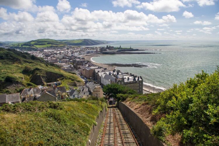 A scenic view looking down the steep tracks of a funicular cliff railway toward the coastal town of Aberystwyth. The sweeping landscape captures the town's terraced buildings, a long curving beach with a promenade, and the sea, all framed by green hillsides under a partly cloudy sky.