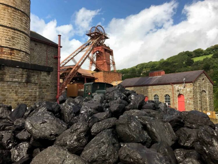 A close-up view of a large pile of rough, black coal in the foreground. Behind it stands the historic mining infrastructure of Rhondda Heritage Park, featuring a tall red metal winding tower, traditional stone buildings with arched windows and red doors, and a vintage green railway carriage. The industrial site is nestled against a backdrop of lush green hills under a partly cloudy blue sky.