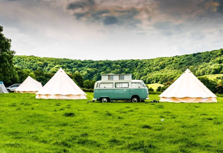 A vintage green camper van is parked on a grassy field between three cream-coloured tents, offering charming glamping in Wales. Surrounded by green hills and trees under a cloudy sky, it’s an idyllic retreat for nature lovers.