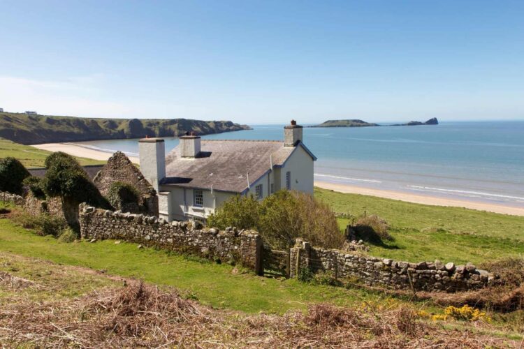 A white cottage with a stone wall overlooks a sandy beach and blue sea, surrounded by green grass and rolling hills under a clear sky—perfect for relaxing farm cottage holidays in Wales