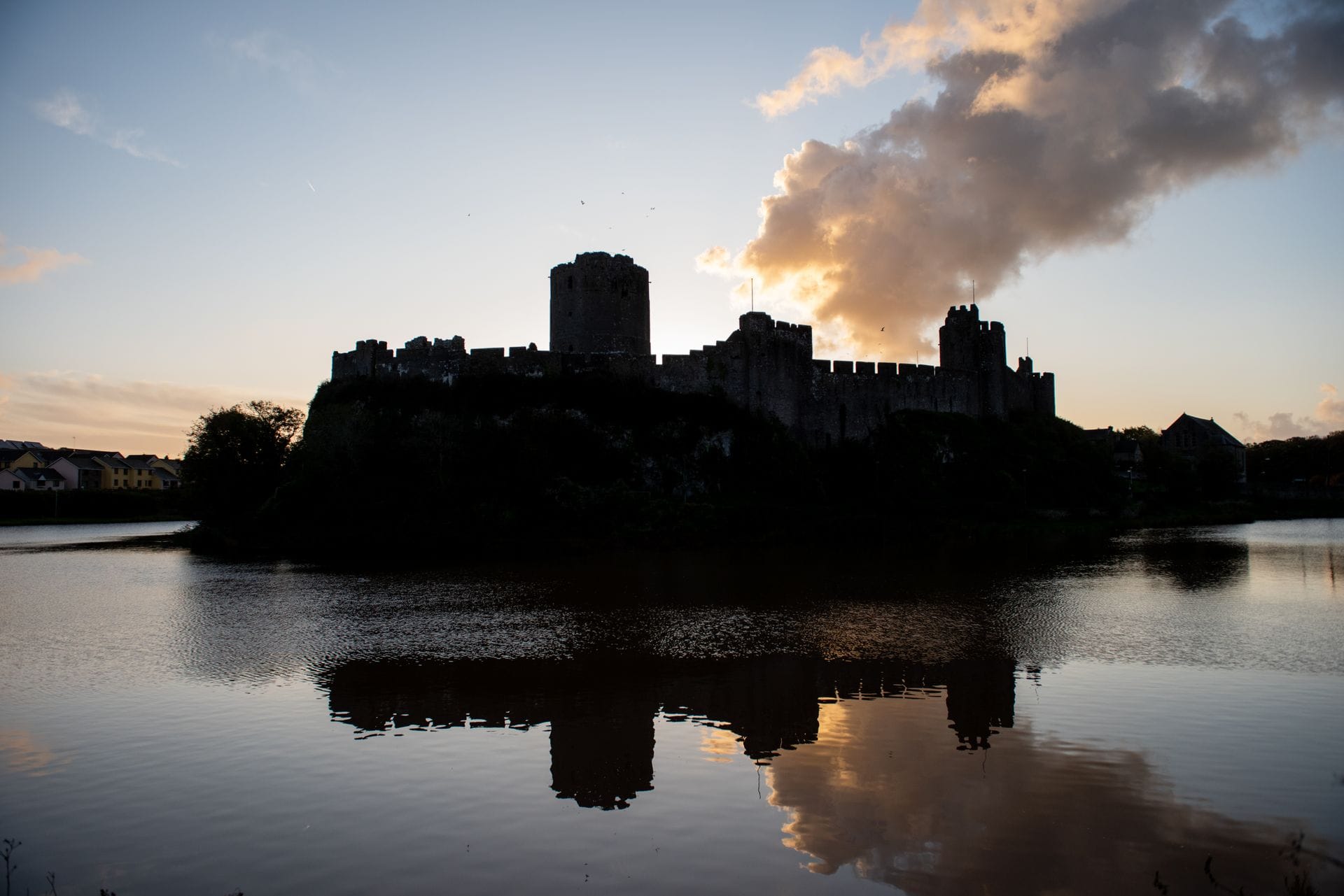 Pembroke Castle silhouetted against a sunset sky, its towers and the Great Keep clearly outlined above the water, with the castle's reflection visible in the still surface below.