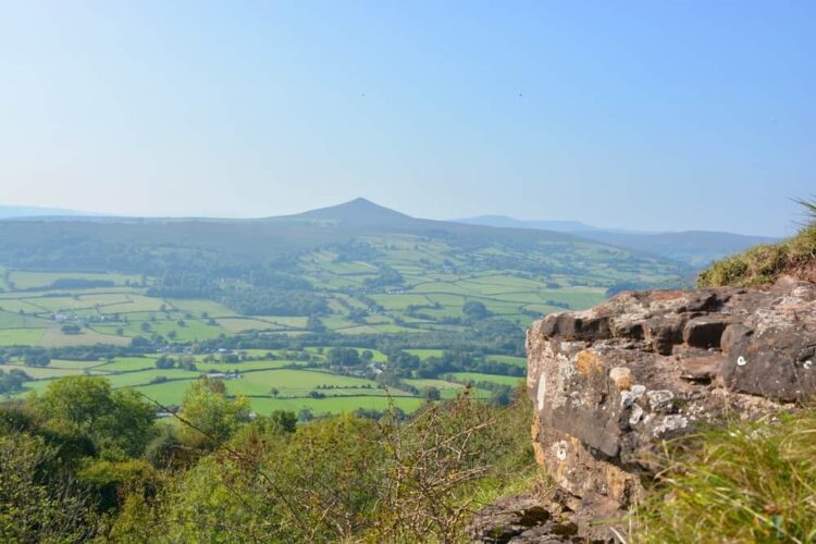 A scenic, panoramic view overlooking the lush green countryside toward the distinctive, pointed peak of the Skirrid mountain near Abergavenny. In the foreground, a rugged stone outcrop sits on a grassy hillside, framing a vast valley of vibrant green patchwork fields and hedgerows under a clear, bright blue sky.