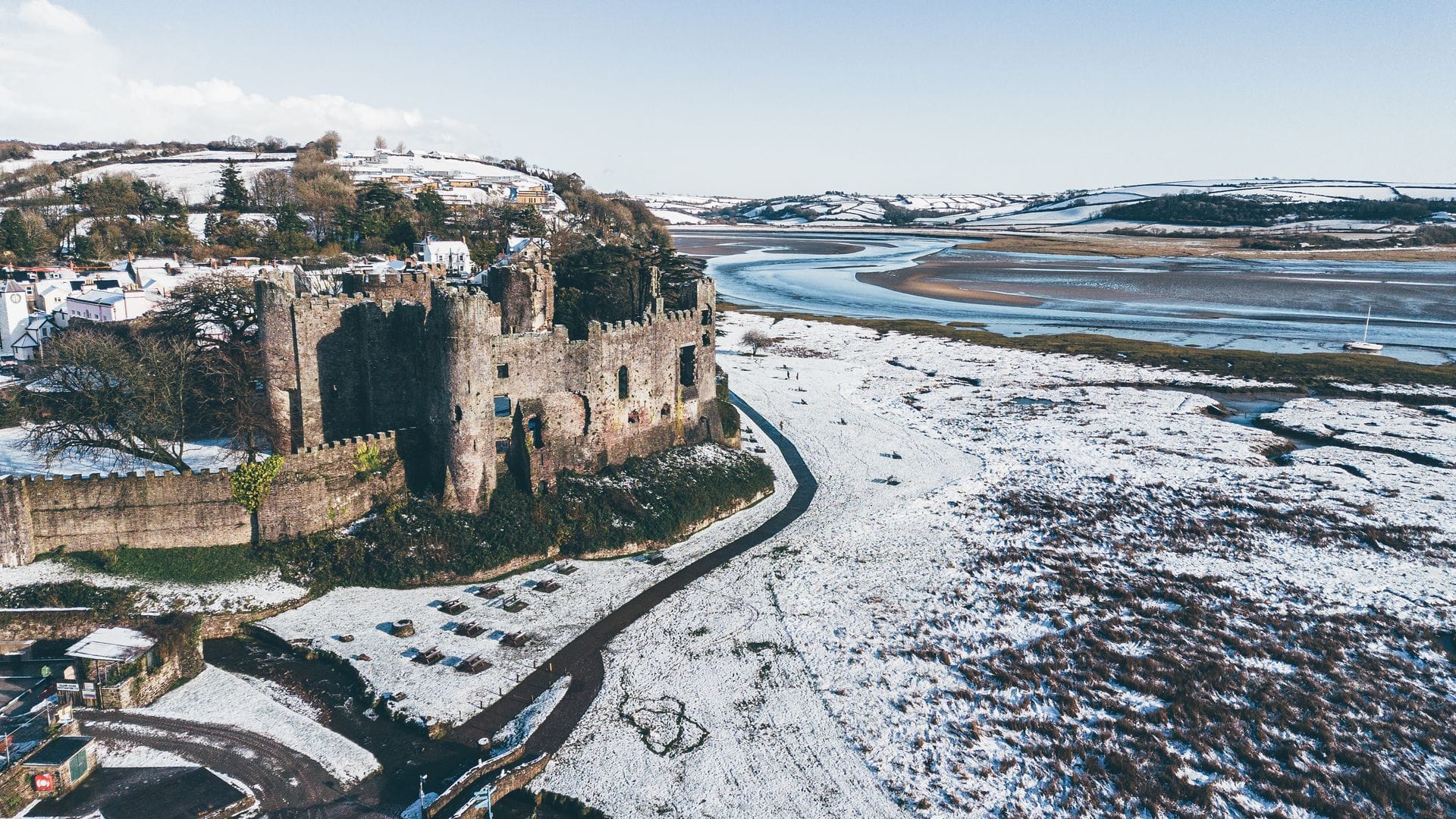 An elevated view of the stone ruins of Laugharne Castle standing on the edge of a wide, winding estuary. The surrounding fields, salt marshes, and distant hills are blanketed in a layer of bright white snow under a clear sky.