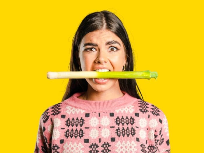 A young woman in a pink patterned jumper holds a leek horizontally in her mouth, looking surprised, against a bright yellow background—one of the quirky things to do in Wales.