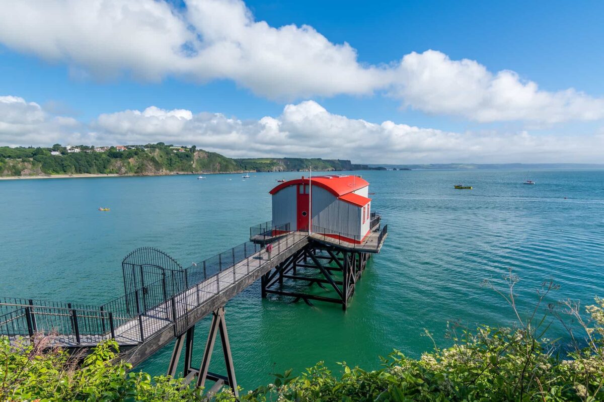A bright sunny view looking down at the converted Old Lifeboat Station in Tenby. The building features grey siding and a curved red roof, sitting on a black stilt structure in the calm blue sea, connected to the lush green cliffs by a long footbridge.