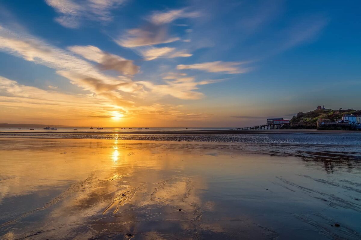 A spectacular golden sunrise reflecting perfectly on the wet sand of North Beach in Tenby at low tide, with the lifeboat station visible in the distance