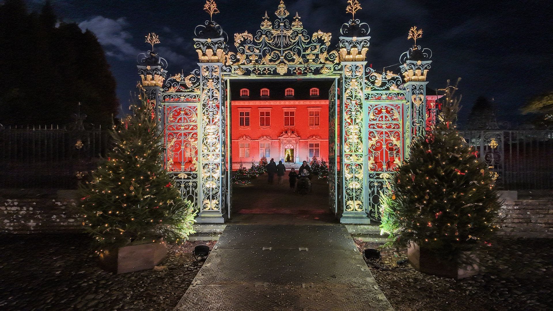 The grand entrance gates of Tredegar House open at night to reveal the historic red-brick mansion illuminated in festive lighting and surrounded by decorated Christmas trees.
