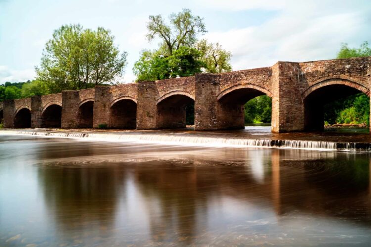 A historic stone bridge with multiple arches spans a gently flowing river, its reflection mirrored in the smooth water—one of the charming sights to discover when you visit Wye Valley Wales, surrounded by lush greenery and tranquil scenery.
