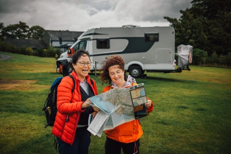 Two women smiling and looking at a map together whilst standing on grass near their Wales campervan, dressed in outdoor jackets with rucksacks, under a cloudy sky.